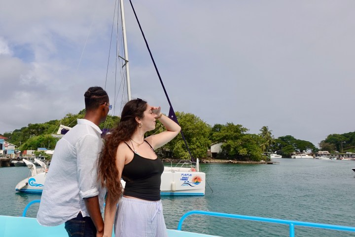 Two people on a boat, one looking out to sea, with other boats and greenery in the background.