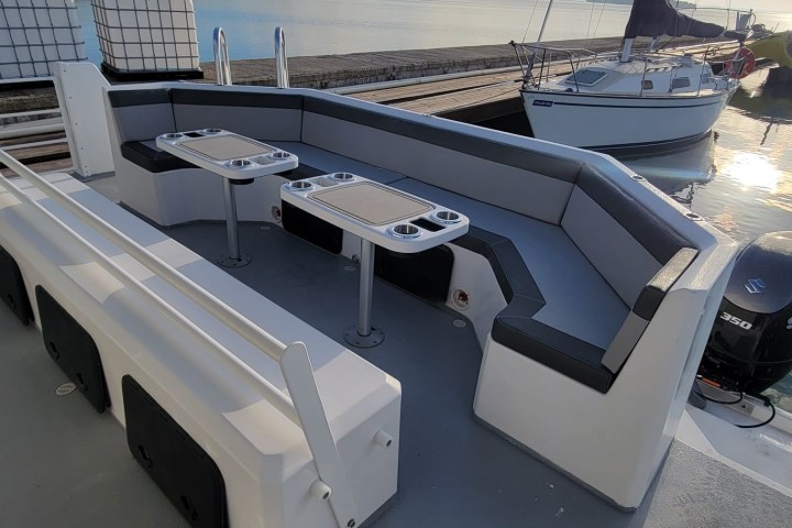 Boat deck with seating and tables, docked by calm water and a sailboat.