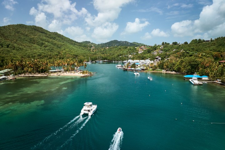 Boats sail on a turquoise bay surrounded by lush green hills and palm trees under a blue sky with clouds.