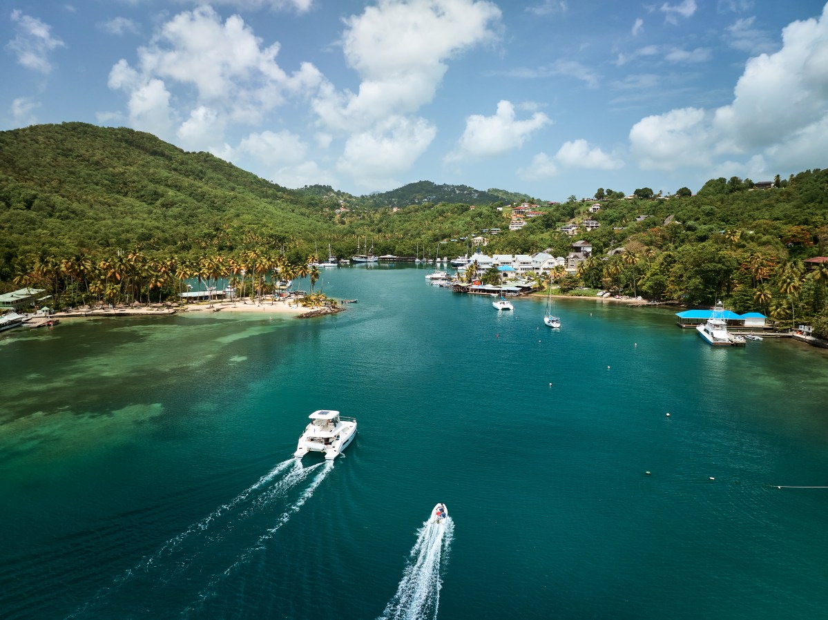 Boats sail on a turquoise bay surrounded by lush green hills and palm trees under a blue sky with clouds.