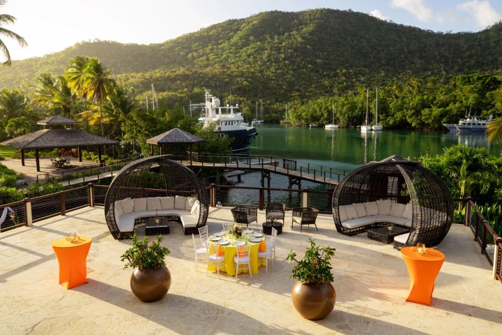 Tropical patio with round sofas, tables, and yachts on a lush, mountainous waterfront.