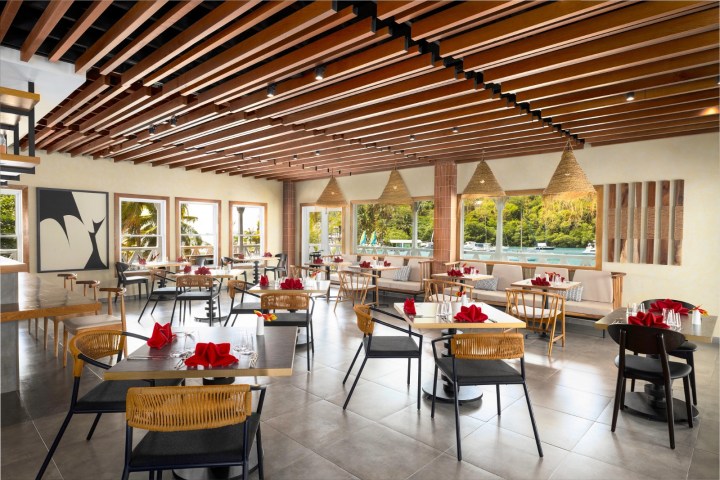 Modern restaurant with wooden beams, tables, red napkins, and large windows overlooking greenery.